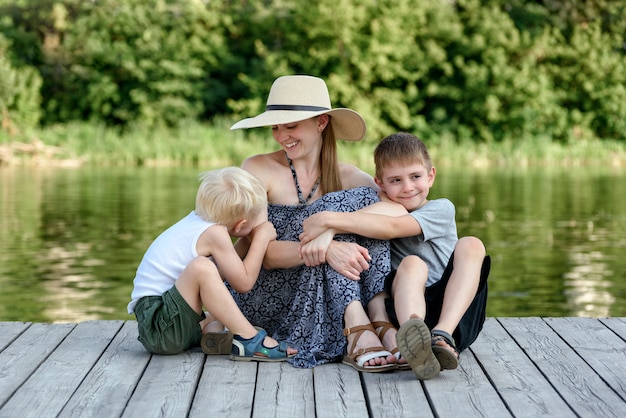 Beautiful mother with two young sons are embracing on the pier on the river bank