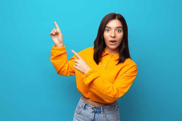 Beautiful multiethnicity woman in orange trendy shirt happily surprised expression pointing up again...