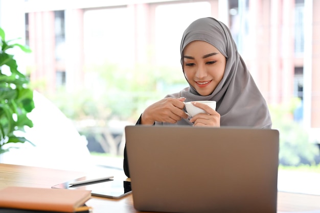 Beautiful Muslim businesswoman with hijab sipping morning coffee looking at laptop screen