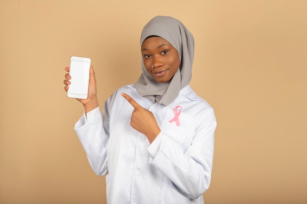 Beautiful muslim religion female doctor with pink bow against cancer shows a blank smartphone screen isolated on a yellow background