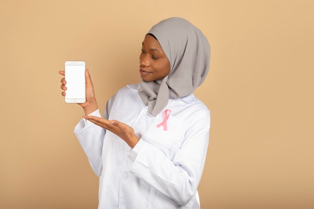 Beautiful muslim religion female doctor with pink bow against cancer shows a blank smartphone screen isolated on a yellow background