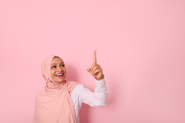 Beautiful Muslim woman of Middle Eastern ethnicity dressed in religious outfit and covered head with hijab ethnicity smiles and looks up pointing her index finger up on a copy space of pink background