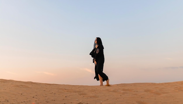 Beautiful mysterious woman in traditional arabic black long dress stands in the desert on sunset