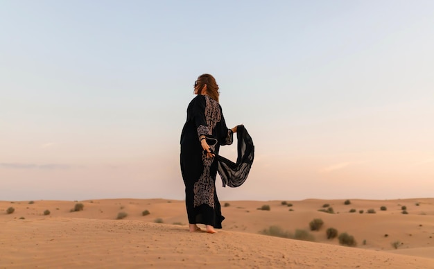 Beautiful mysterious woman in traditional arabic black long dress stands in the desert on sunset