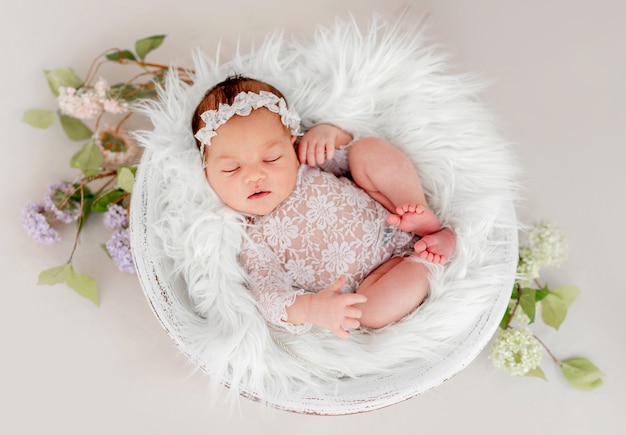 Beautiful newborn baby girl wearing lace costume ana wreath sleeping in basin filled white fur during studio photoshoot. Cute portrait of infant child napping