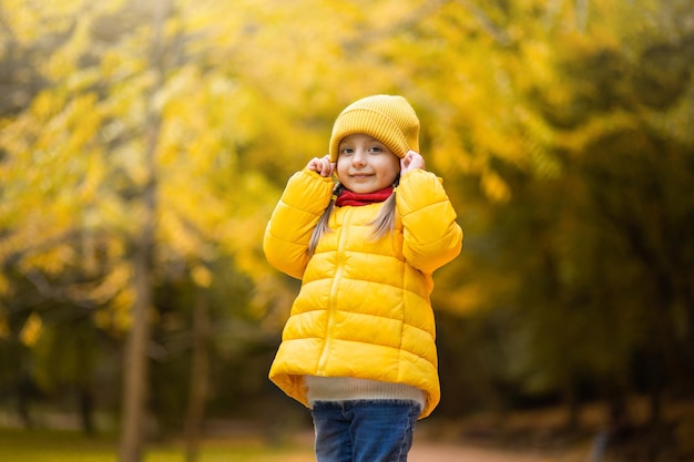 Beautiful outdoor autumn portrait of adorable 4-years old child girl, wearing stylish yellow cap and coat, touching her cap and smiling at camera