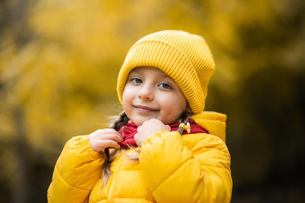 Beautiful outdoor autumn portrait of adorable 4-years old child girl, wearing stylish yellow cap and coat, touching her warm red scarf and smiling at camera.