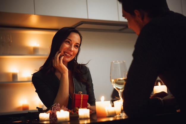 Beautiful passionate couple having a romantic candlelight dinner at home, drinking wine, woman getting a gift box