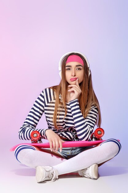 Beautiful pensive woman sitting with skateboard and listening to music on pink background