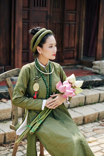 Beautiful pensive young Vietnamese woman in ao dai dress sitting on wooden stool with lotus flowers