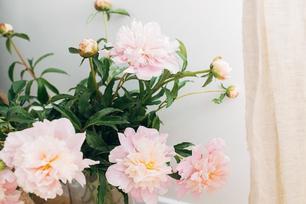 Beautiful peonies bouquet in modern boho room Gentle pink peony flowers in vase on rustic background moody image Modern bohemian decor stylish comfy interior details