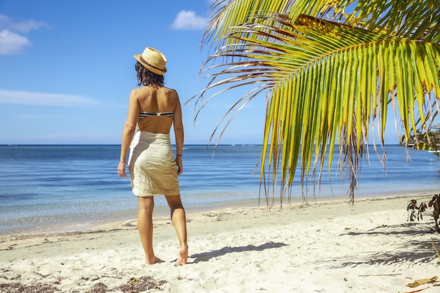 Beautiful picture of a brunette female in a swimsuit next to a palm against blue lagoon at daytime