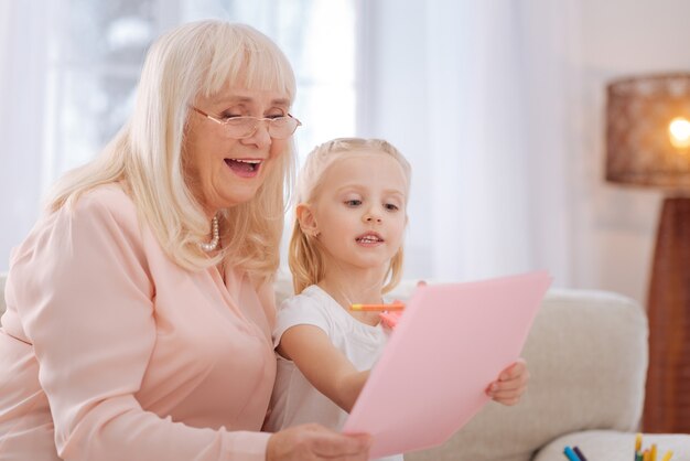 Beautiful picture. Nice pleasant aged woman sitting together with her granddaughter and holding a drawing while looking at it