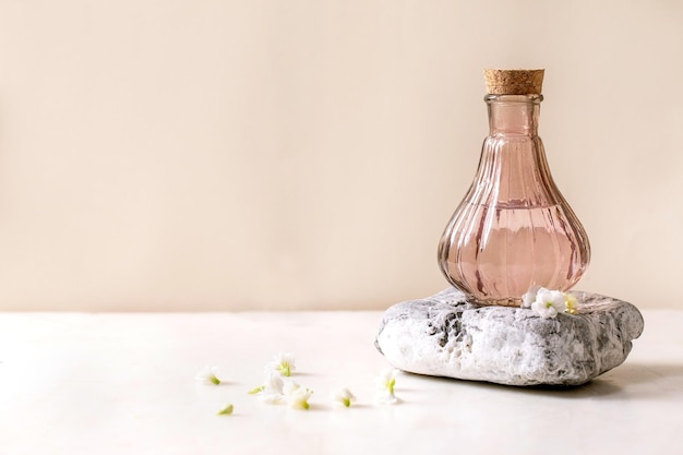 Beautiful pink transparent bottle of perfume with cork standing on textured stone on white marble table with small white flowers Kalanchoe around Perfume making concept with place for design label