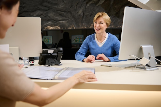 Beautiful pleasant Caucasian female travel agent talking to her client showing catalog with tours or consumer services while sitting in modern office