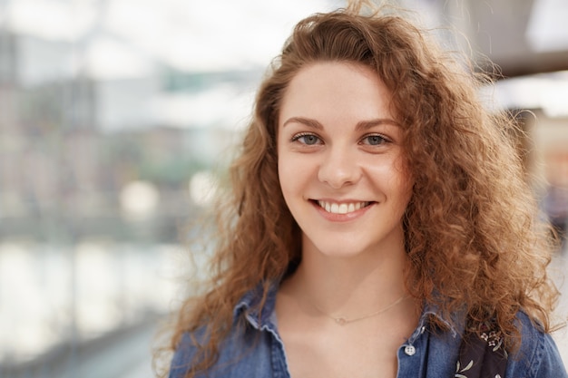 Beautiful pleased female with cheerful expression, dressed in fashionable clothing, poses indoor, recreats in shopping centre