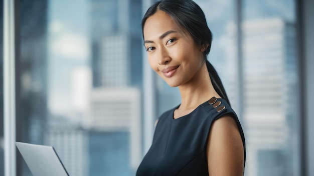 Photo beautiful portrait of an asian businesswoman in stylish black dress using laptop computer posing next to window in city office confident female ceo smiling successful diverse business manager