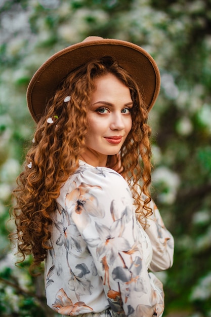 Beautiful portrait of an attractive young girl with blossom of an apple tree in her curly hair. Female wearing beige hat and white dress in the spring park. New season concept.