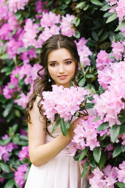 Beautiful portrait of a lovely girl who stands surrounded by lilac pink flowers blooming rhododendron in the spring in the garden