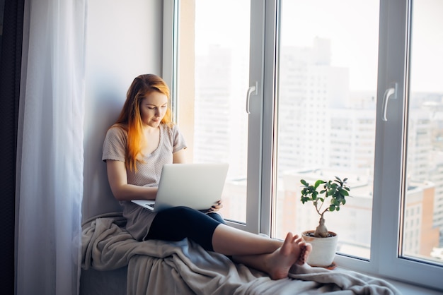 Beautiful positive girl using laptop, sitting on the windowsill in city apartment. Young red-haired woman working at home. Freelance concept.