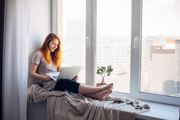 Beautiful positive girl using laptop, sitting on the windowsill in city apartment. Young red-haired woman working at home. Freelance concept.