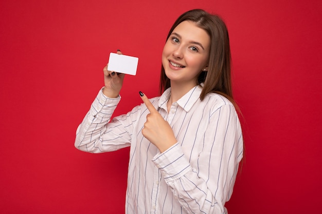 Beautiful positive smiling young dark blonde woman wearing white blouse isolated over red background holding credit card looking at camera pointing finger at plastic contactless card.