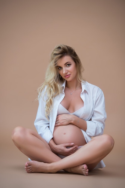 beautiful pregnant blonde woman sits in a white shirt and underwear on a beige wall