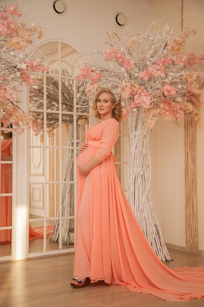 A beautiful pregnant girl in a dress and a beige interior hugs her belly