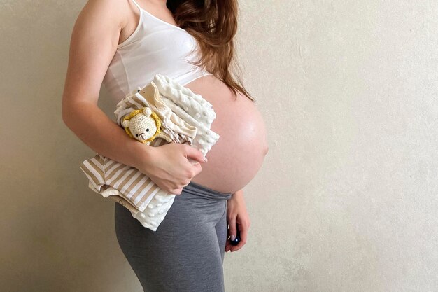 A beautiful pregnant girl is holding a stack of clothes for a newborn Preparation for the appearance of the baby Children's shopping Copy space