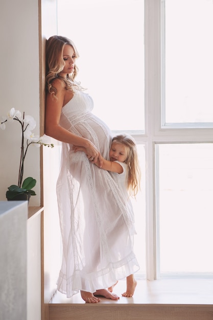 Beautiful pregnant mom with child posing near window at home