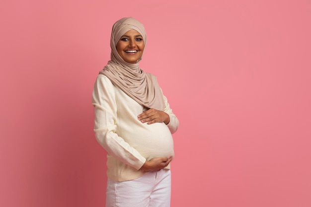 Beautiful Pregnant Muslim Woman In Hijab Embracing Belly And Looking At Camera