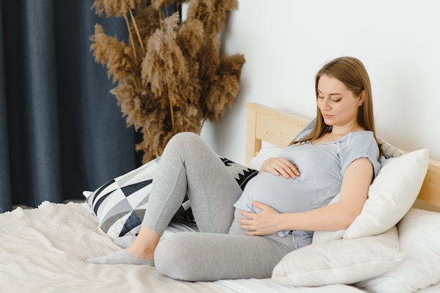 Beautiful pregnant woman holds hands on belly in bedroom at home. Young mother waiting of a baby. Concept of pregnancy, maternity, health care, gynecology, medicine. Close-up, indoors.