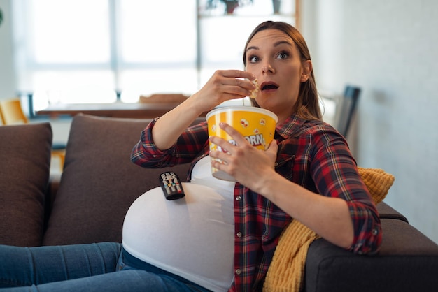 Beautiful pregnant woman is eating popcorn, looking at camera and smiling while sitting on couch