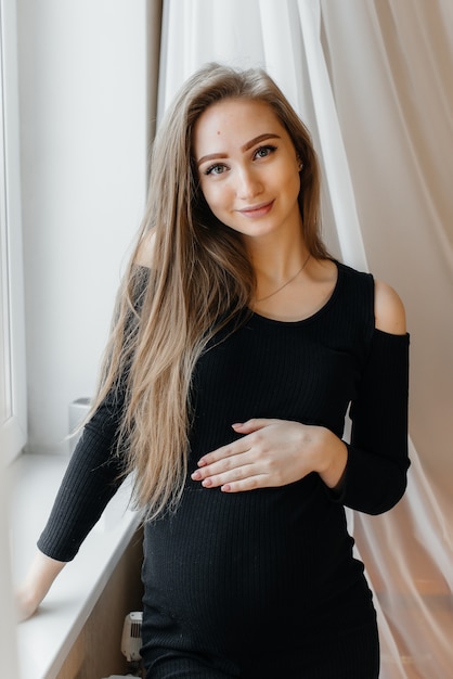 A beautiful pregnant woman is standing in a room near the window