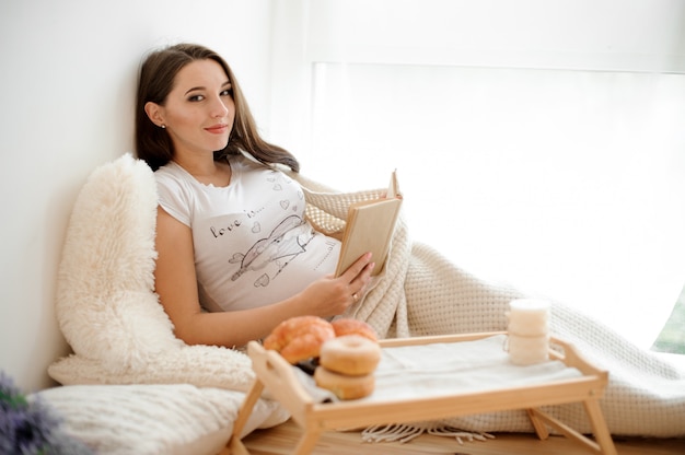 Beautiful pregnant woman lying on the white bed with a book