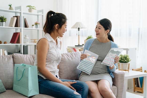 Beautiful pregnant woman open gift box from best friend and smiling while sitting on couch among shopping bags at home. girl send give baby clothes as present in baby shower. motherhood pregnancy.