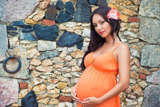 Beautiful pregnant woman in an orange sundress at the stone wall background. Old village Altos de Chavon, Dominican Republic