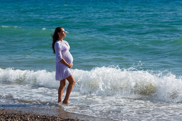 Beautiful pregnant woman standing on beach in light dress in the wind Pleasure