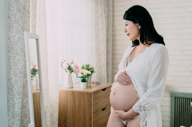 Beautiful pregnant woman with bare belly looking mirror in light room. young future motherhood smiling checking reflection with her unborn baby inside naked tummy. sexy childbearing mom in lingerie