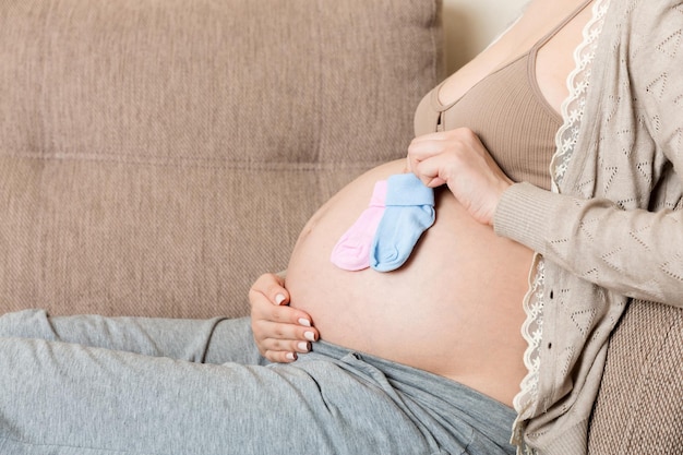 Beautiful pregnant young woman holding a pair of cute baby socks on her tummy lying on sofa at home