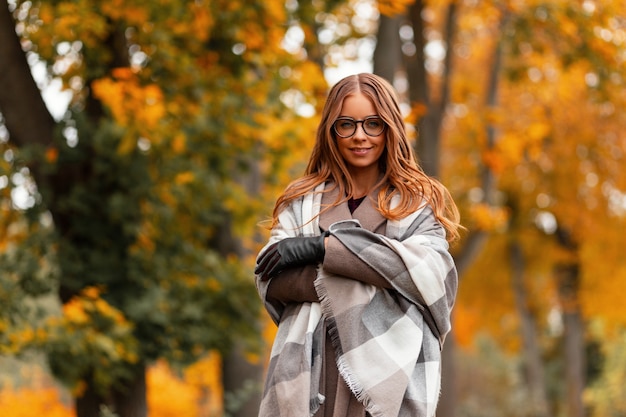 Beautiful pretty happy woman model with glasses in a coat in a knitted scarf enjoys relaxing in the autumn park among a tree with orange foliage. Cheerful girl stands in the park and smiles cute.