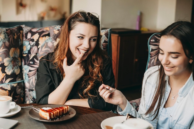 Beautiful red hair woman laughing while tasting a cake with her finger while looking at her girlfriend sitting in a coffee shop.