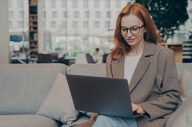 Beautiful red-haired business woman in casual clothes and eyeglasses working online, using laptop, sitting on comfortable grey couch in office lounge, female entrepreneur enjoying distant work