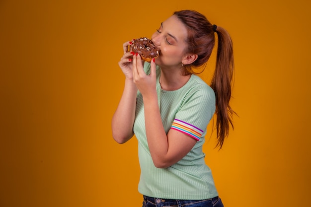 Photo beautiful red-haired girl eating chocolate donuts on yellow background. girl eating donuts