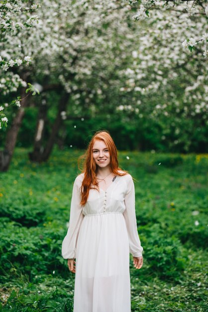 Beautiful red-haired girl in a white dress among blossoming apple-trees in the garden.