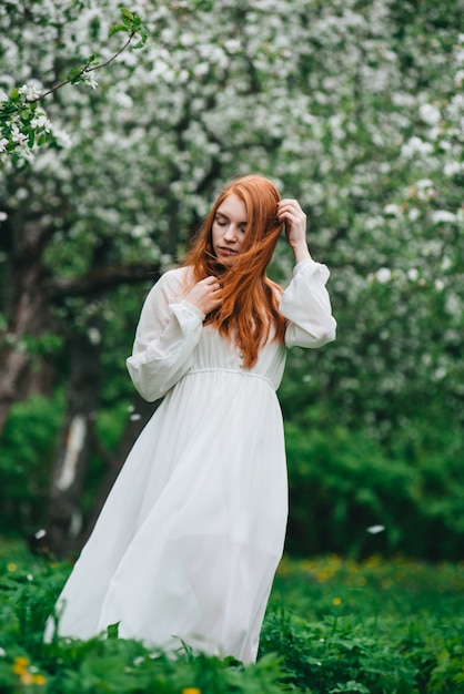 Beautiful red-haired girl in a white dress among blossoming apple-trees in the garden.