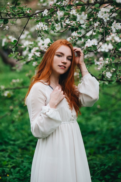 Beautiful red-haired girl in a white dress among blossoming apple trees in the garden 