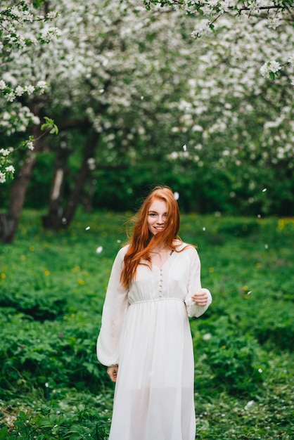 Beautiful red-haired girl in a white dress among blossoming apple trees in the garden 