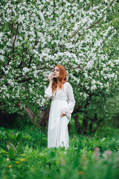 Beautiful red-haired girl in a white dress among blossoming apple trees in the garden 