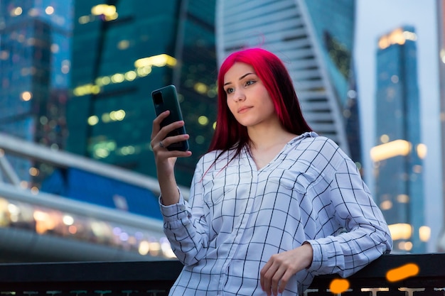 Beautiful red-haired girl with smartphone in the evening on lighted city street.